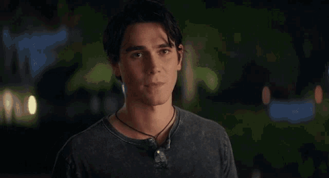 a young man wearing a black t-shirt and a necklace is looking at the camera at night .