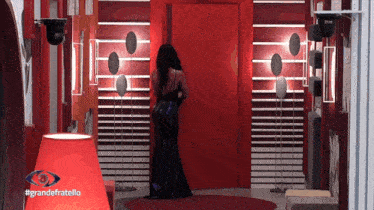 a woman in a long black dress is standing in front of a red wall with the words grandefratello written on it