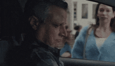 a man is sitting in the driver 's seat of a car looking out the window at a woman .