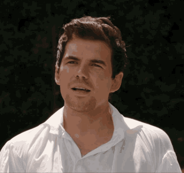 a man in a white shirt looks at the camera with trees in the background