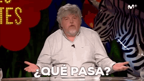 a man with a beard is sitting at a table with a zebra in the background and says " que pasa "