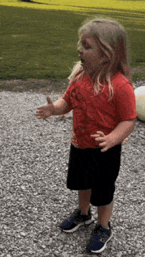 a little girl wearing a red shirt and black shorts is standing on a gravel road