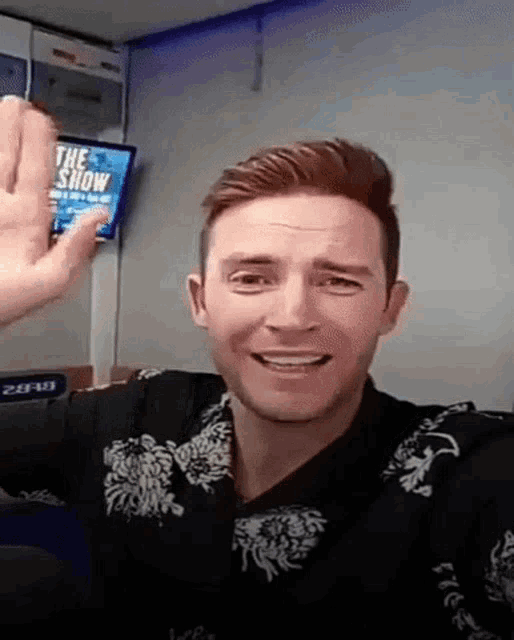 a man is smiling and waving at the camera while wearing a floral shirt .