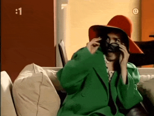 a woman in a red hat and green coat is sitting on a couch talking on a cell phone .