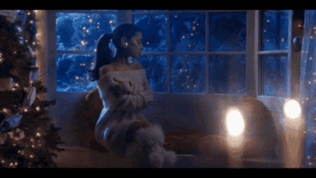 a woman is sitting on a window sill in front of a christmas tree and looking out the window .