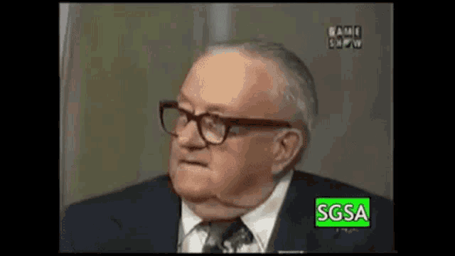 an older man wearing glasses and a suit is sitting in front of a television .