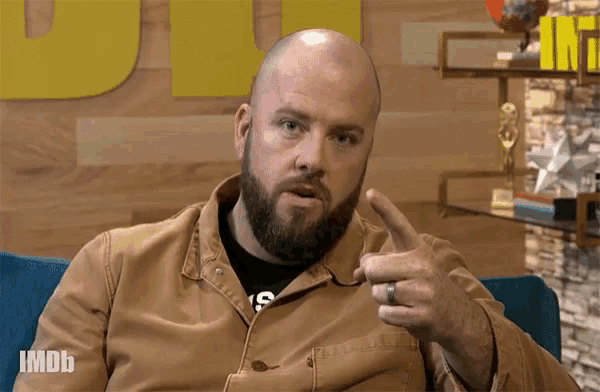 a bald man with a beard is sitting in a chair with imdb written on the bottom of his jacket