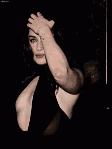 a woman in a black dress with a plunging neckline holds her hand to her head