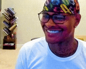 a man wearing glasses and a bandana smiles in front of a book shelf