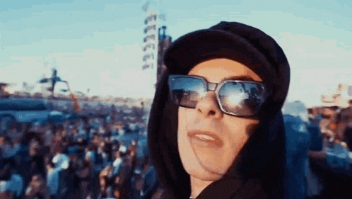 a man wearing sunglasses and a hooded jacket is standing in front of a crowd .
