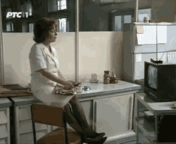 a woman sits at a desk in front of a tv with ptc 1 written on the wall behind her