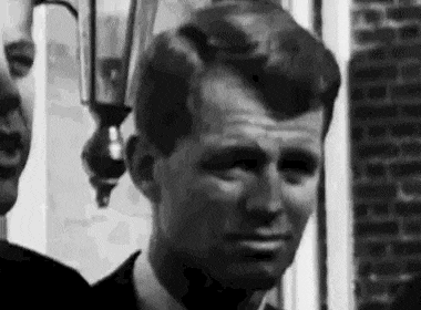 a black and white photo of a man in a suit standing next to another man in front of a brick building .