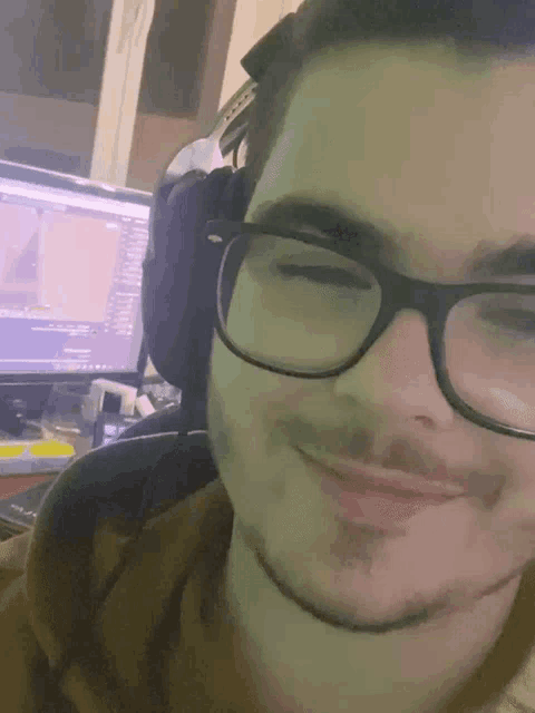 a man wearing glasses and headphones smiles in front of a computer monitor