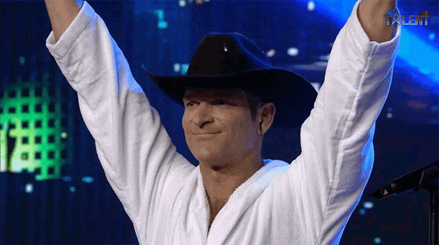 a man wearing a cowboy hat and a white robe holds his arms up in the air