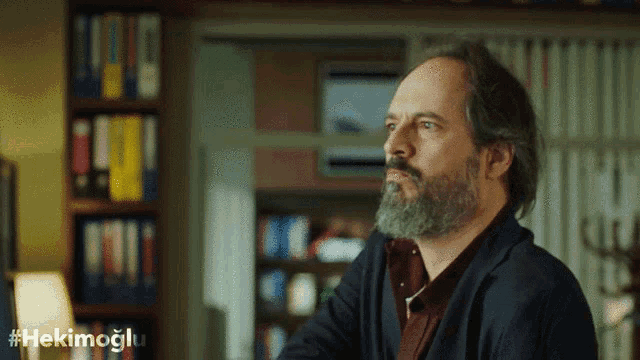 a man with a beard is sitting in front of a bookshelf with the hashtag #hekimoglu