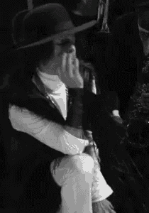 a black and white photo of a man wearing a hat covering his face with his hand .