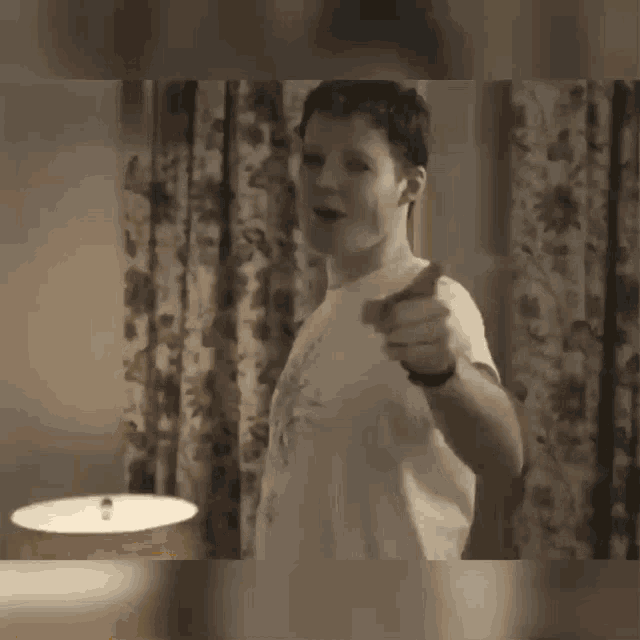 a man in a white shirt is pointing at the camera while standing in front of a window .