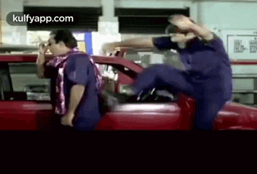 two men are standing next to a red car and one of them is kicking the other .