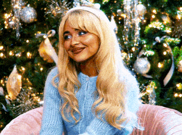 a blonde woman wearing a blue sweater is sitting in front of a christmas tree .
