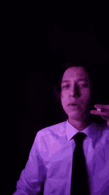 a man in a white shirt and black tie is smoking a cigarette in a dark room .