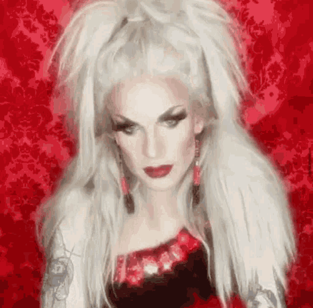 a drag queen with long white hair and red lipstick is standing in front of a red background .