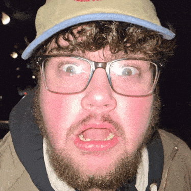 a man with a beard wearing glasses and a hat making a face