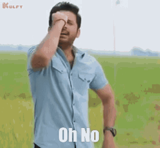 a man in a blue shirt is standing in a field and covering his face with his hand and saying oh no .