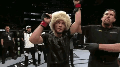 a man wearing a furry hat holds up his hand in a boxing ring