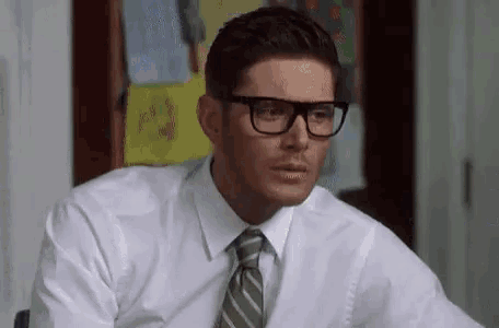 a man wearing glasses , a white shirt , and a tie is sitting at a desk .
