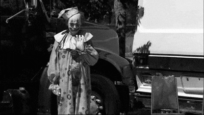 a black and white photo of a clown standing in front of a dumpster