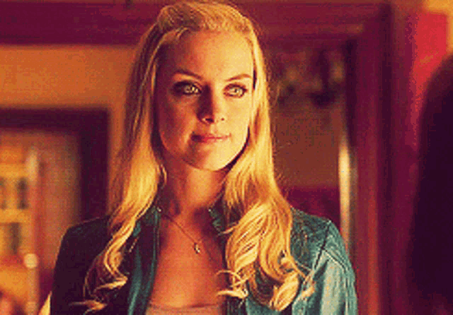 a blonde woman wearing a blue jacket and a necklace looks at the camera