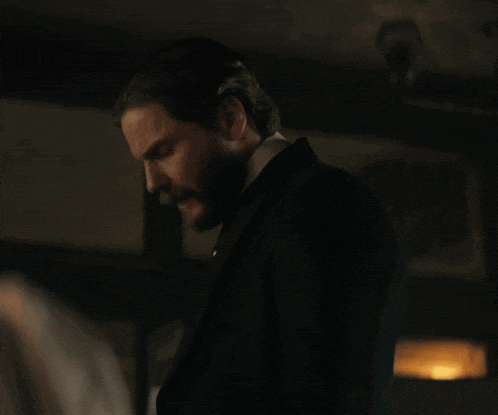 a man with a beard wearing a suit and tie stands in a dark room