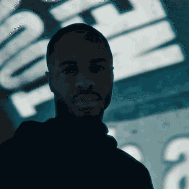 a man with a beard looks at the camera in front of a blurred sign that says ' e '
