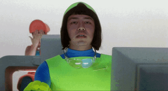 a man wearing a green vest and goggles stands in front of a computer