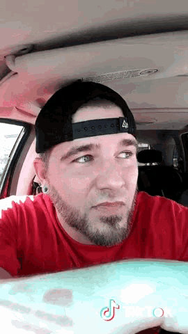 a man wearing a red shirt and a black hat is sitting in a car with tiktok written on the bottom right