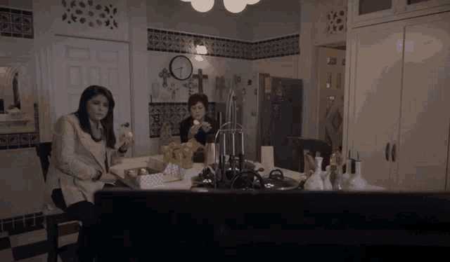 two women sitting at a table in a kitchen with a clock on the wall