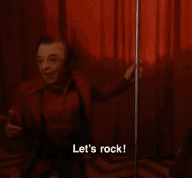 a man in a red shirt is standing in front of a red curtain and says let 's rock