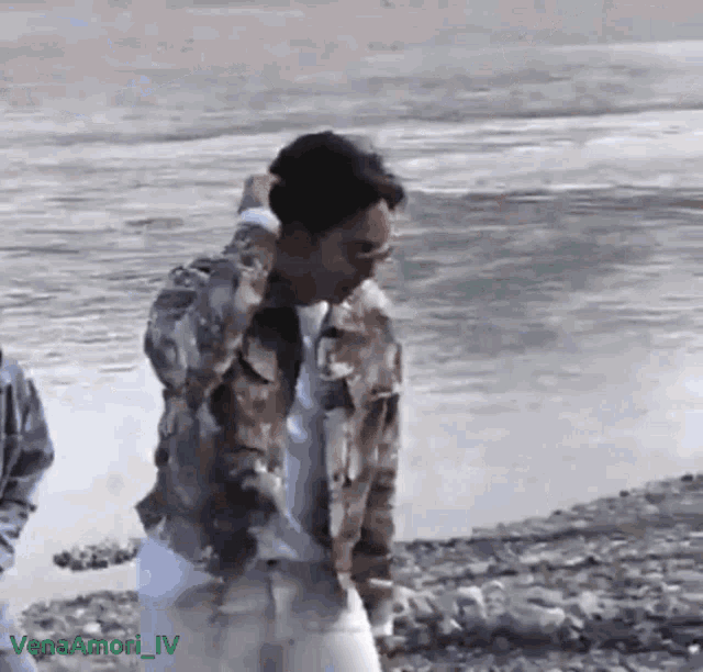 a man in a camouflage jacket is standing on a rocky beach near a body of water ..