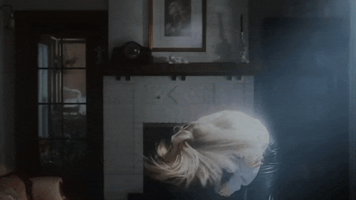 a woman is standing in front of a fireplace in a living room and her hair is blowing in the wind .