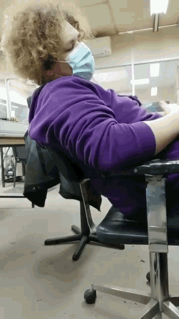 a woman wearing a purple sweatshirt and a blue face mask is sitting in a chair