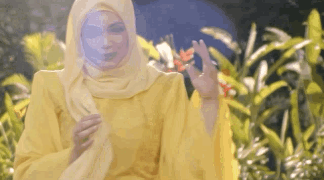 a woman wearing a hijab and a yellow dress is holding a flower in her hand .