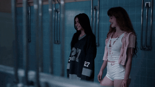 two girls are standing in a bathroom with one wearing a jersey with the number 27 on it
