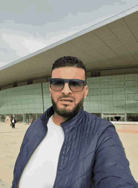 a man wearing sunglasses and a blue jacket takes a selfie in front of a building
