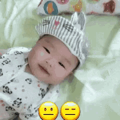 a baby wearing a hat with bunny ears is laying on a bed with two smiley faces next to him .