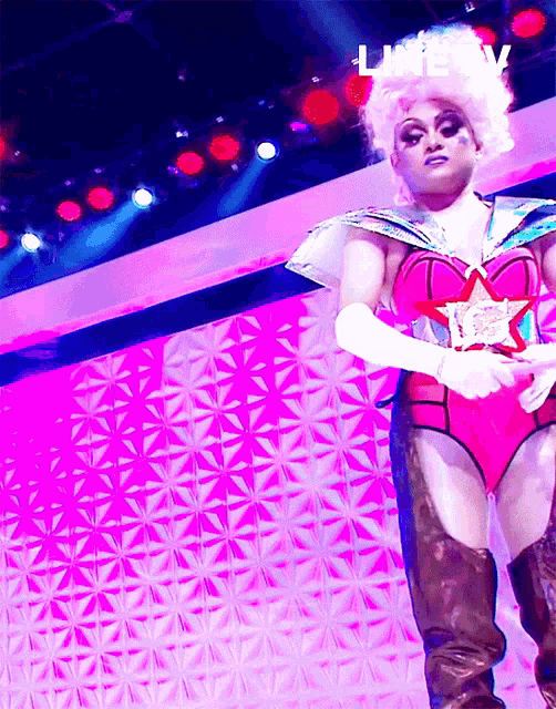 a drag queen stands on a stage in front of a pink wall that says ' live tv '