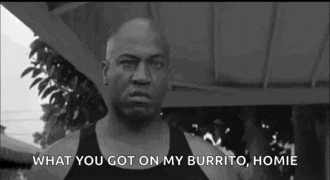 a man in a black tank top is standing in front of a building and talking about a burrito .