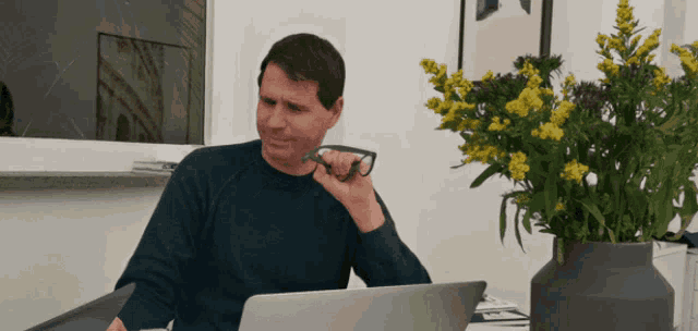 a man sitting at a desk with a laptop and a vase of yellow flowers