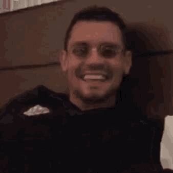 a man wearing sunglasses and a black shirt is smiling and sitting on a bed .