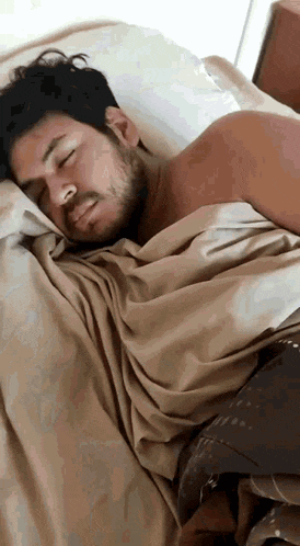a man with a beard is sleeping in a bed with a brown blanket .