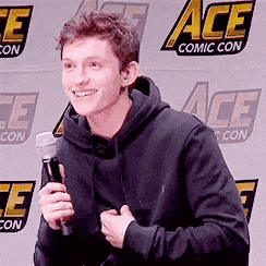 a young man is holding a microphone and smiling while standing in front of a wall with ace comic con written on it .
